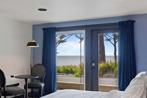 Habitació d'hotel amb un llit, una cadira, una taula, una làmpada i vista a l'oceà a través d'una porta de vidre.