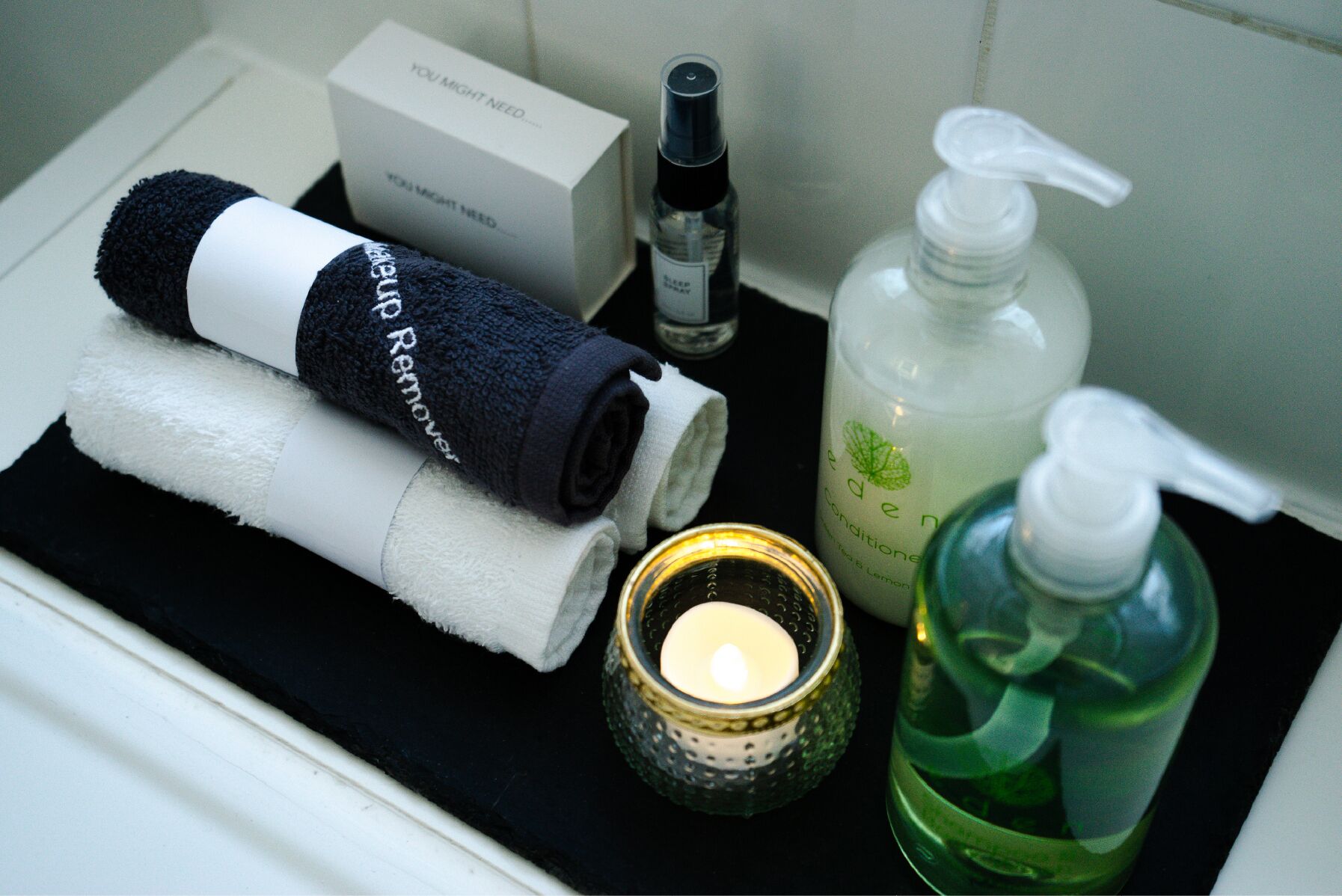 Un bagno con un asciugamano nero, un asciugamano bianco, una scatola con l'etichetta 'Potresti aver bisogno', una bottiglia di spray per dormire, una bottiglia di sapone liquido verde, un erogatore di sapone verde e una candela accesa.