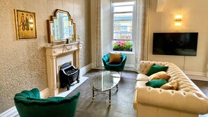 Un salon avec une cheminée, des fauteuils verts, un canapé beige et un téléviseur à écran plat.