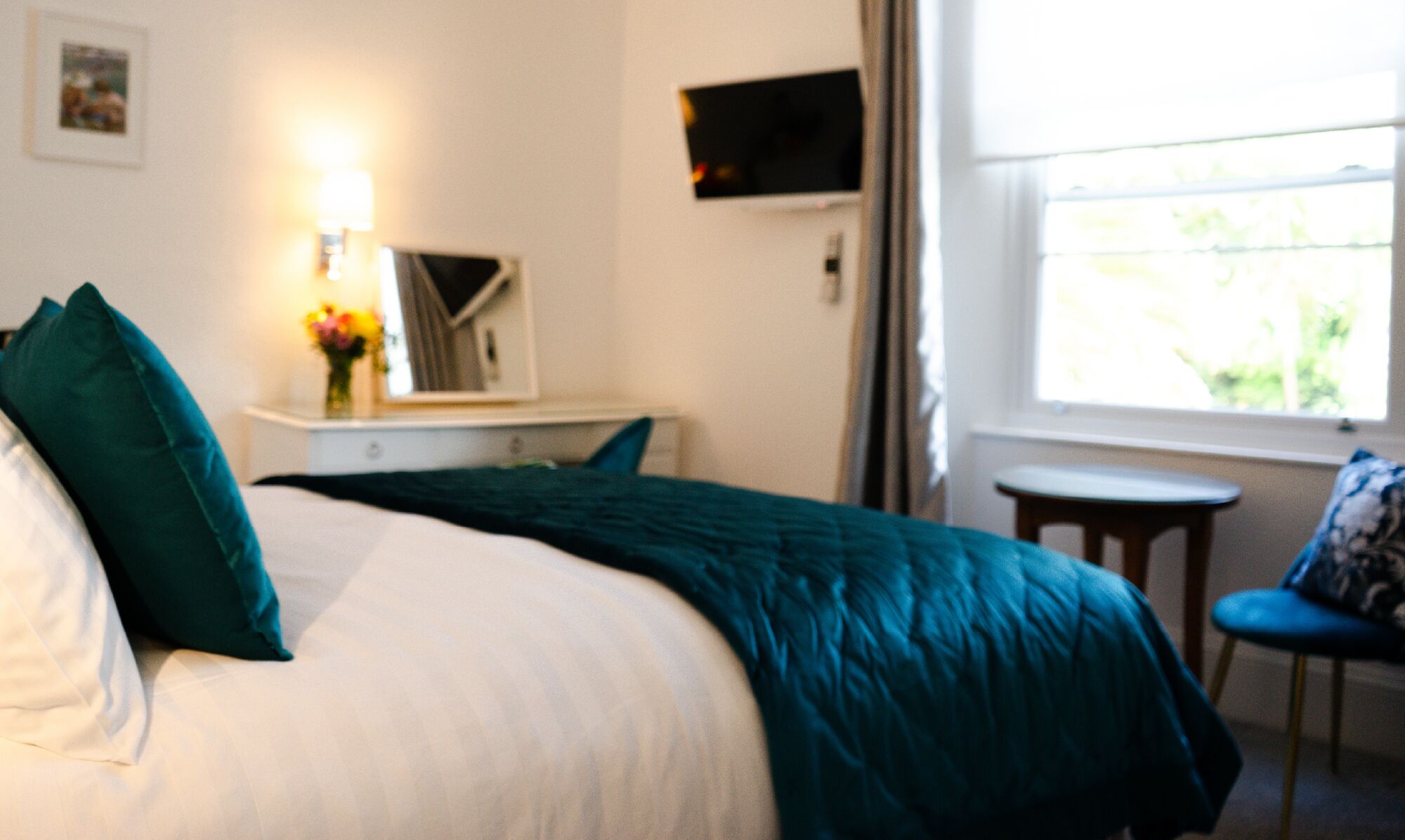 Ein Hotelzimmer mit einem Bett, einem kleinen Tisch mit Stuhl, einem an der Wand befestigten Fernseher und einem Fenster mit Blick auf Grünflächen.