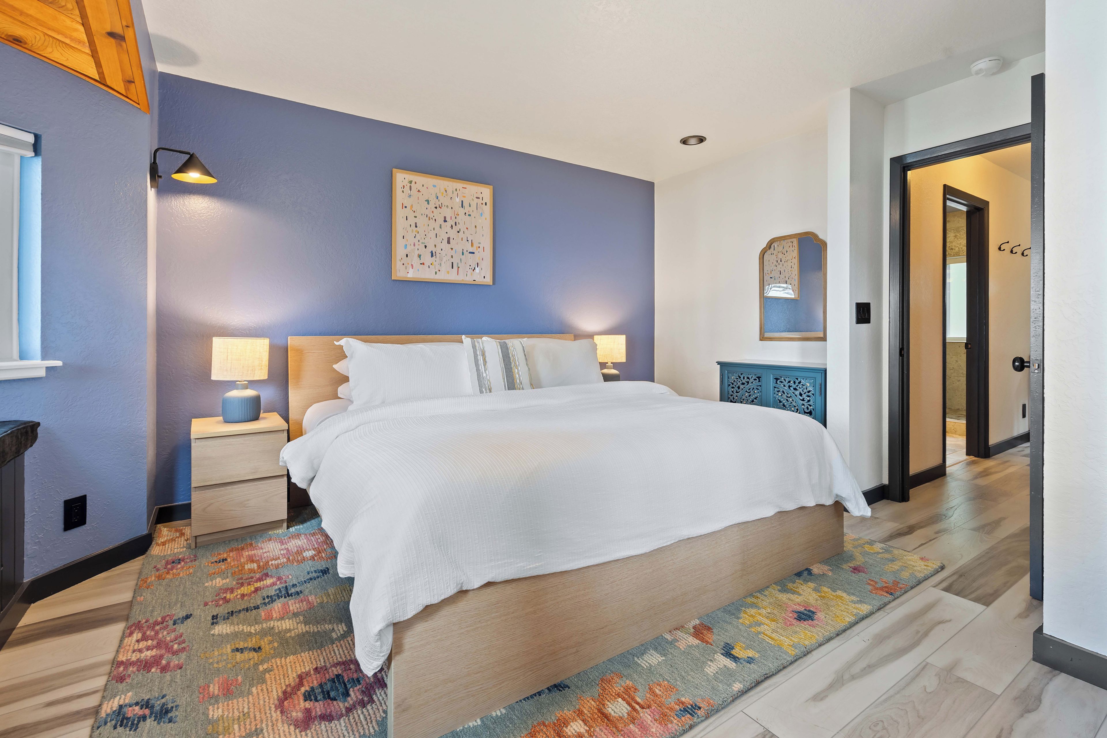 A bedroom with a large bed, a nightstand, a lamp, a wall art piece, and a door leading to another room.