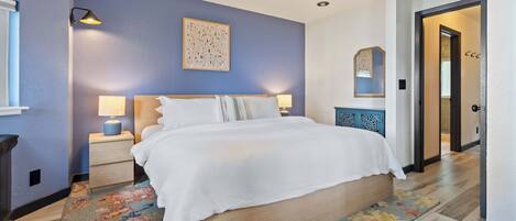 A bedroom with a large bed, a nightstand, a lamp, a wall art piece, and a door leading to another room.