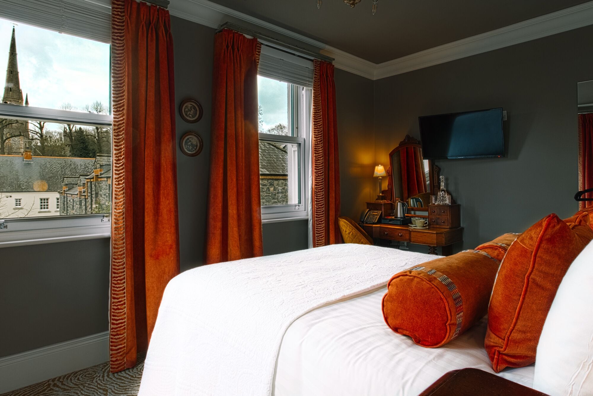 Habitación de hotel con una cama grande, un escritorio con lámpara, televisión y vistas a la aguja de una iglesia por la ventana.