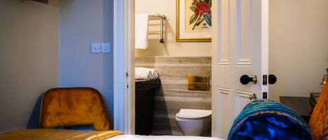 Une chambre d’hôtel avec un lit, une chaise, une salle de bain comprenant des toilettes et un lavabo, ainsi qu’un tableau encadré au mur.