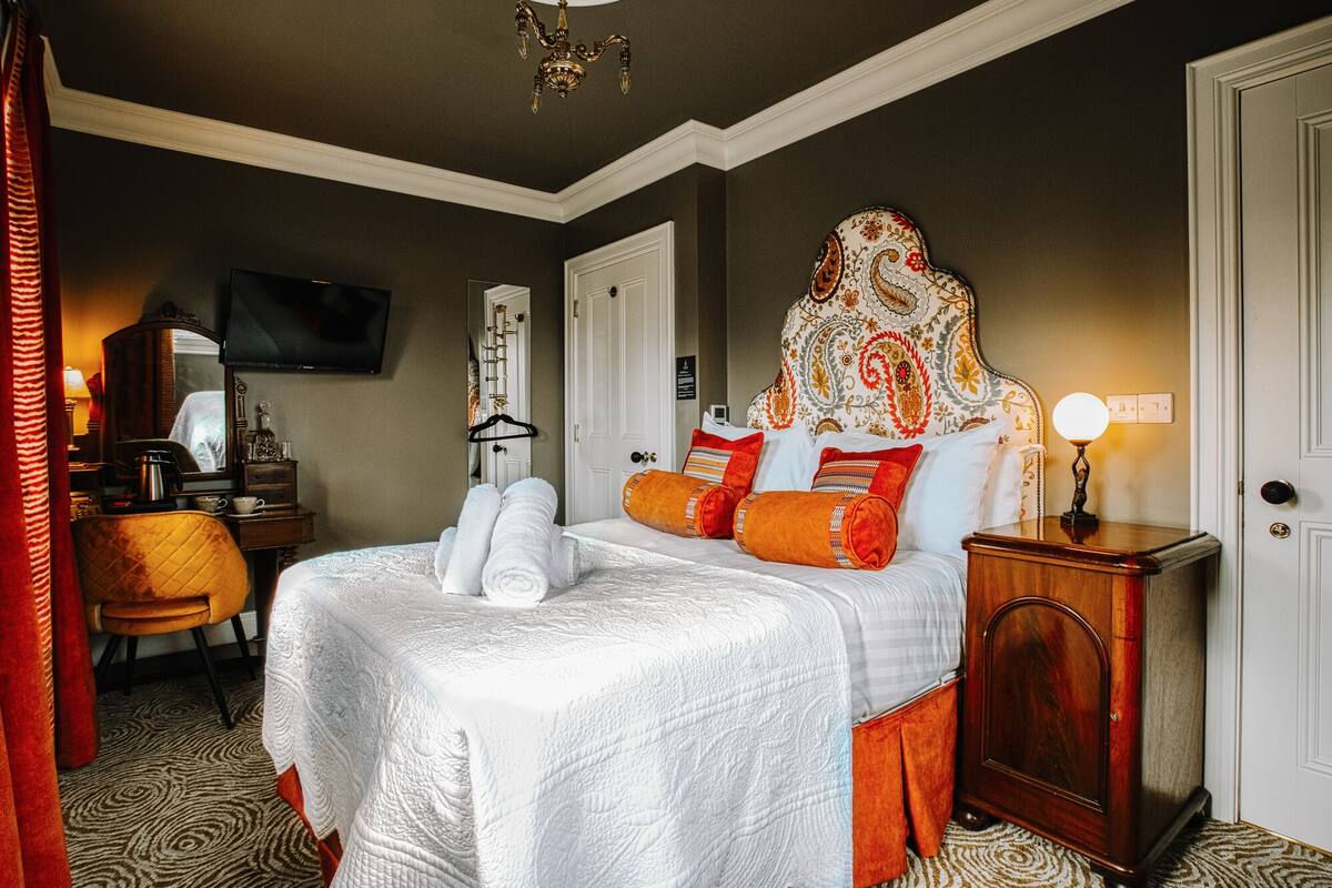 A hotel room with a large bed, a wooden nightstand, a TV, a desk with a chair, and a patterned headboard.