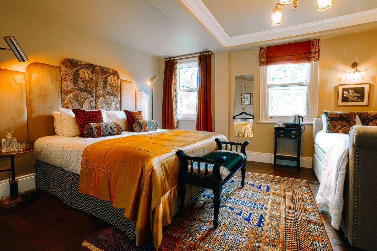A well-lit bedroom with a large bed, a small table with a kettle, and a chair.