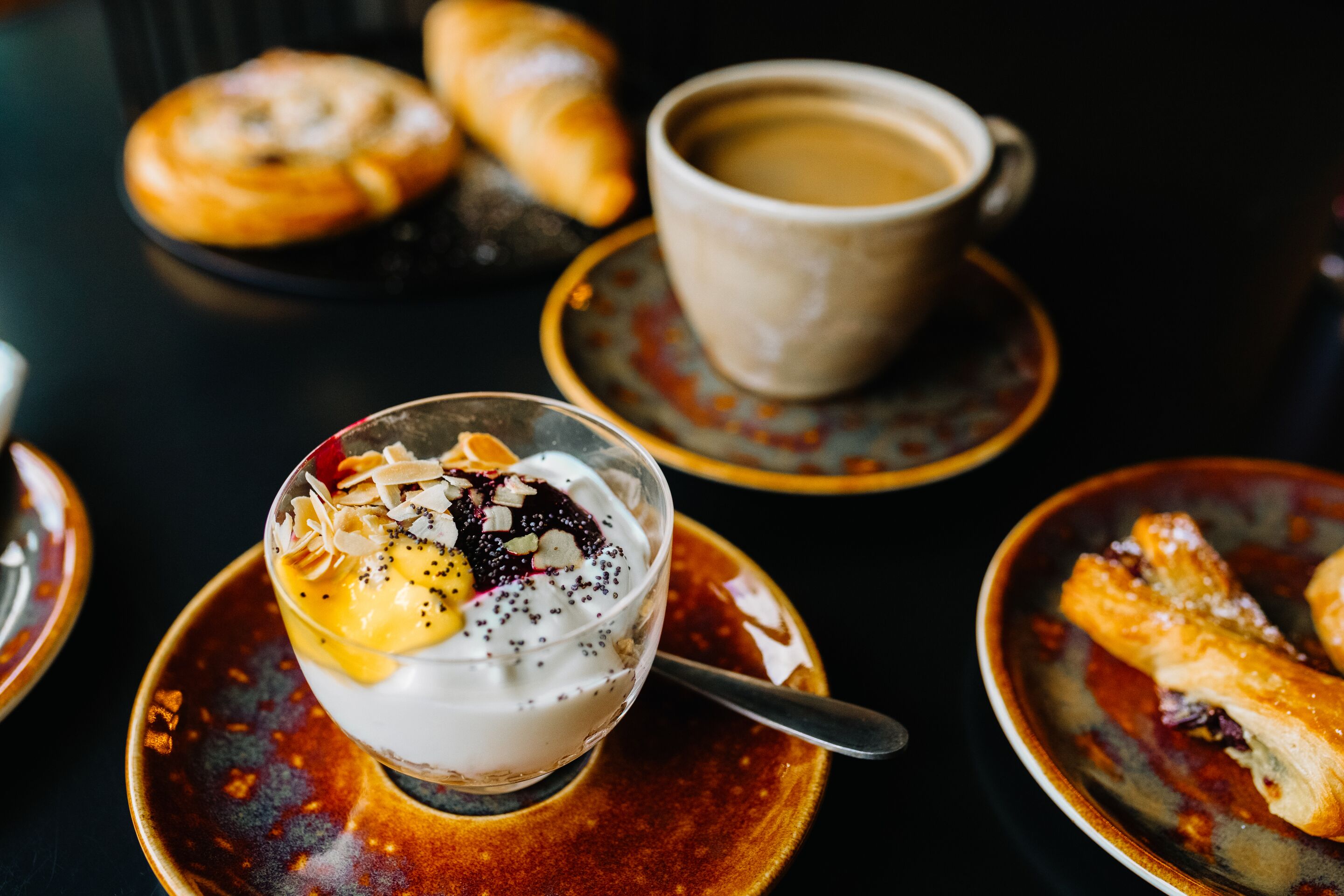 Un postre con yogur, fruta y semillas, una taza de café y pasteles sobre una mesa.