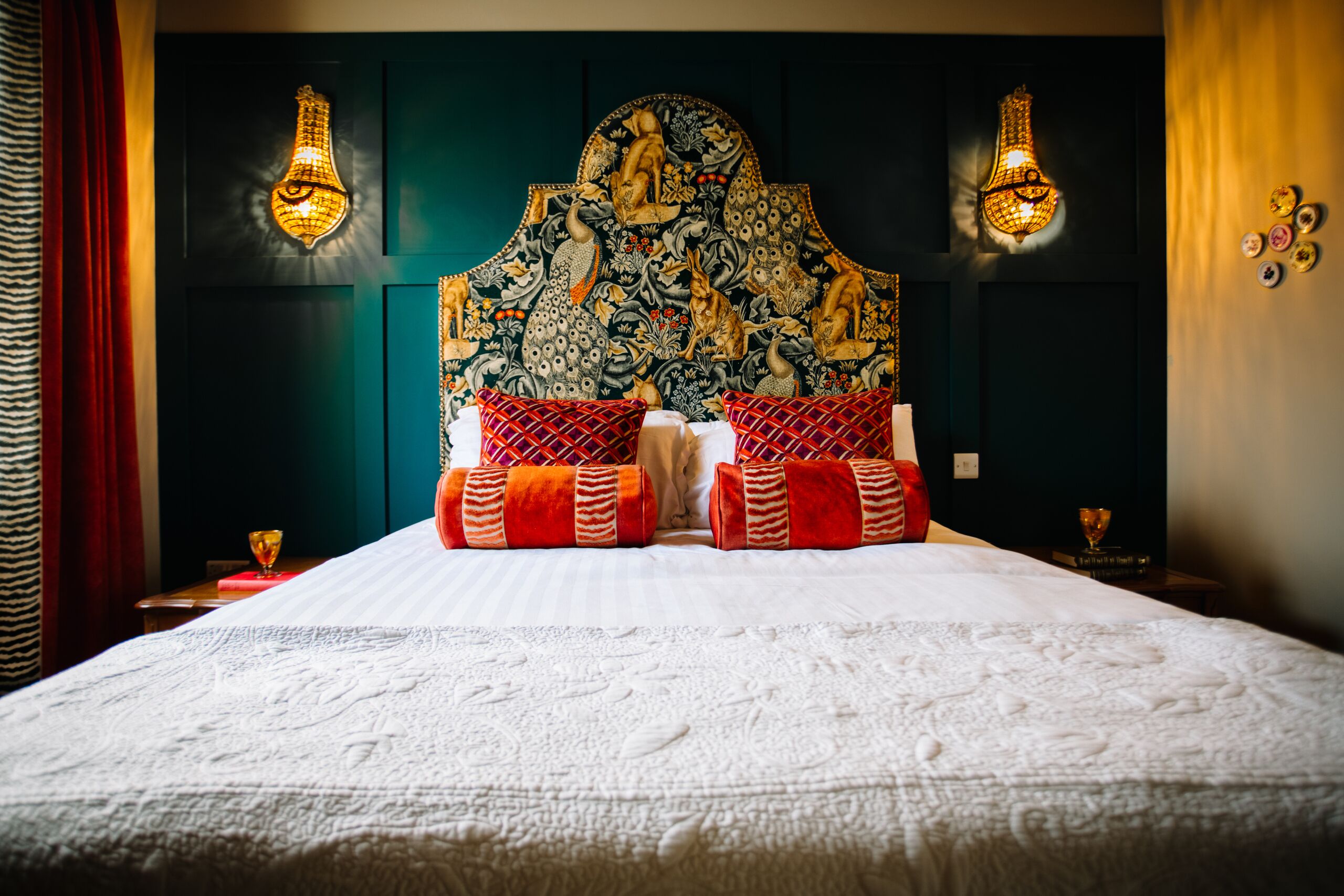 Una cama con un cabecero decorativo, dos apliques de pared y una almohada estampada en rojo y blanco.