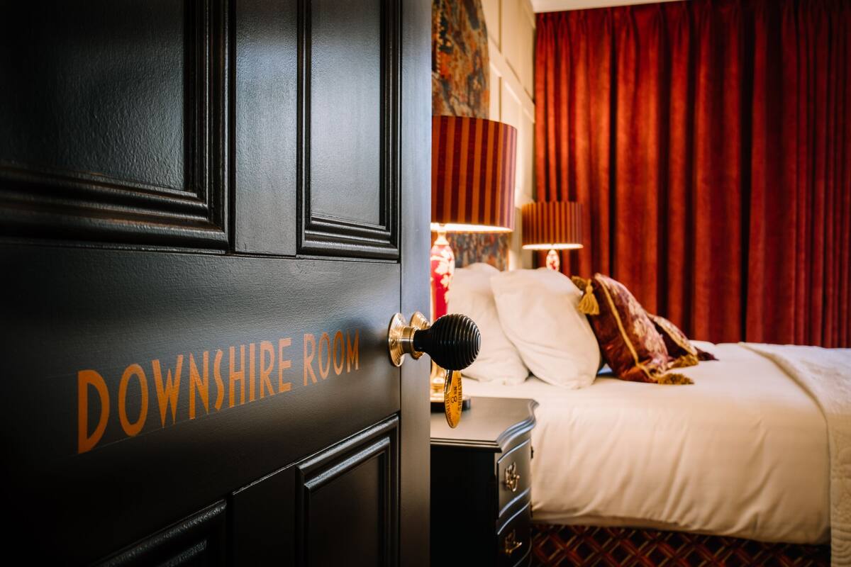 A hotel room with a bed, a nightstand, a lamp, and a door labeled "DOWNSHIRE ROOM".