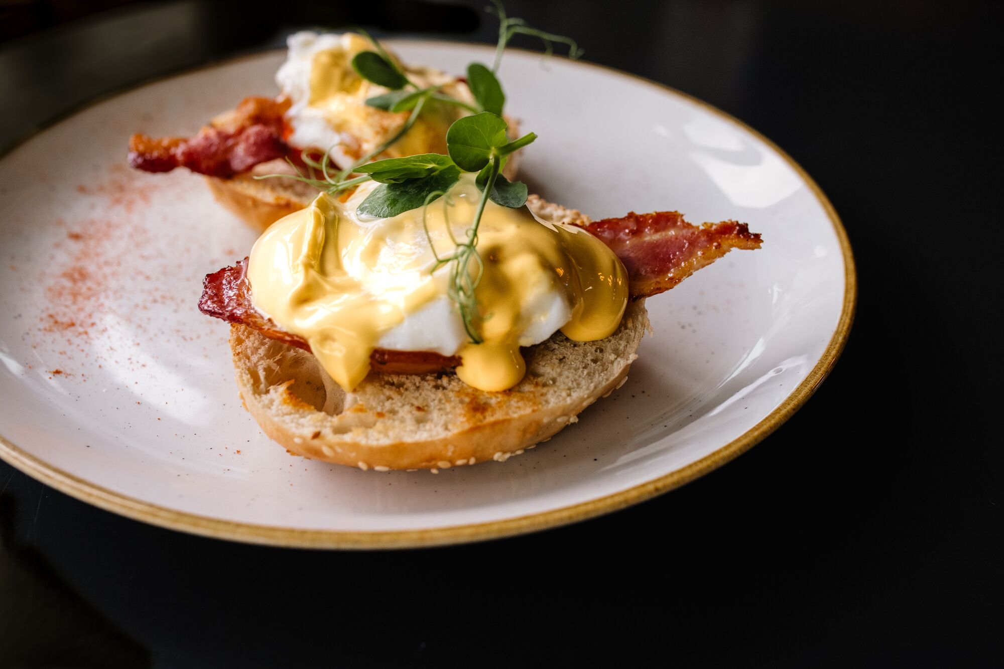 A plate with a breakfast dish consisting of a poached egg, hollandaise sauce, bacon, and a garnish of microgreens.