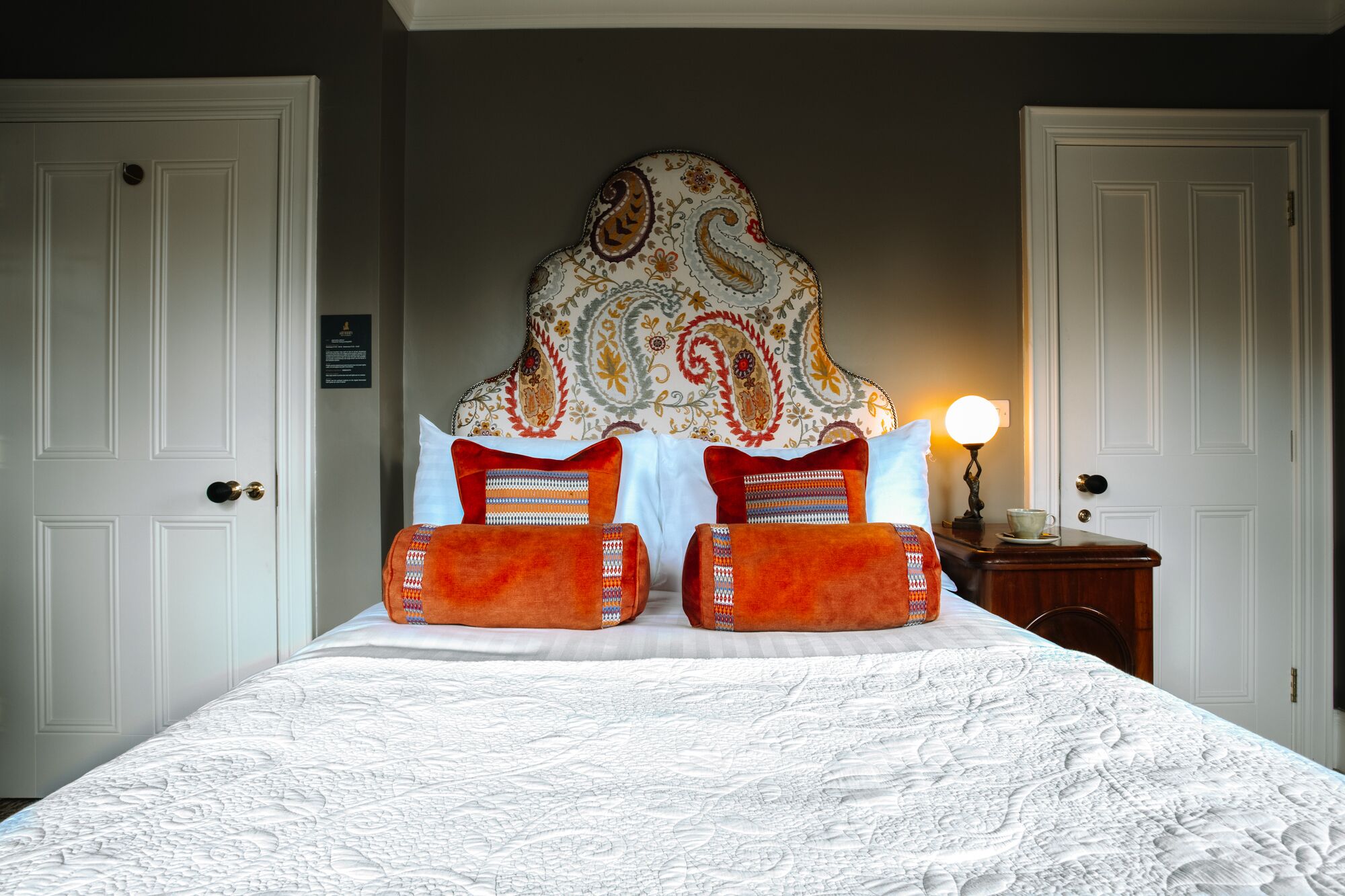 A neatly made bed with a patterned headboard, white walls, and a nightstand with a lamp.