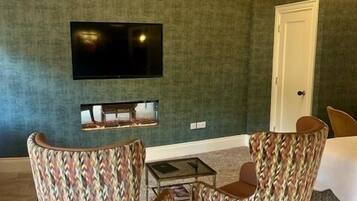 Um quarto com televisão na parede, duas poltronas estampadas, uma mesa pequena e uma porta.