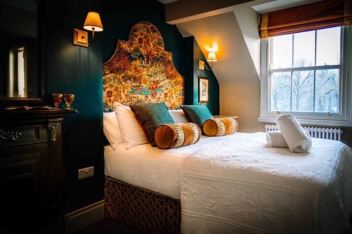 A bedroom with a large bed, a headboard with a floral pattern, two wall sconces, and a window with a view of trees.