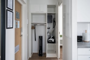 Een smalle gang met een houten deur, een zwarte stofzuiger en een witte opslagunit met planken.