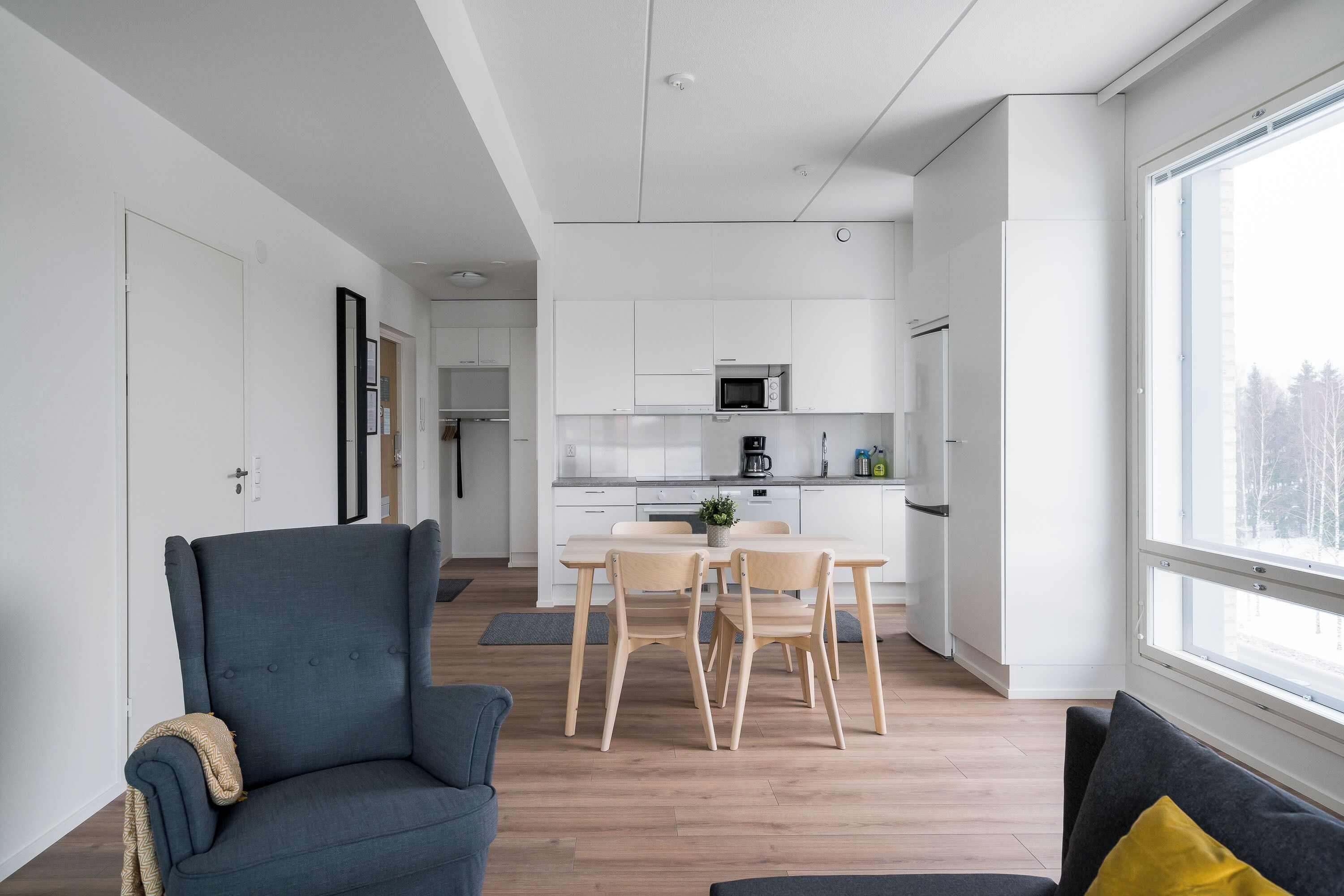 A modern kitchen with white cabinets, a microwave, and a coffee maker. There is a dining table with chairs and a sofa in the living area.