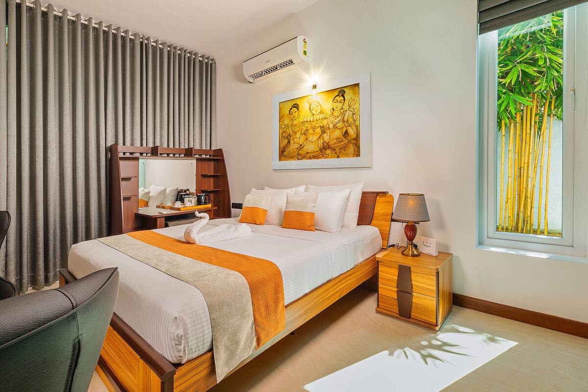 A hotel room with a bed, a desk, a chair, a window with a view of bamboo, and an air conditioning unit.