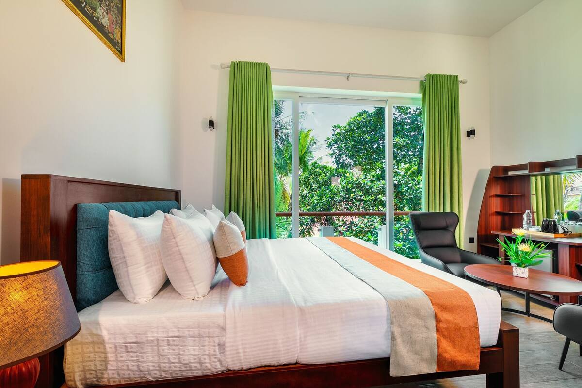 A hotel room with a bed, a desk, a chair, and a view of greenery through the window.
