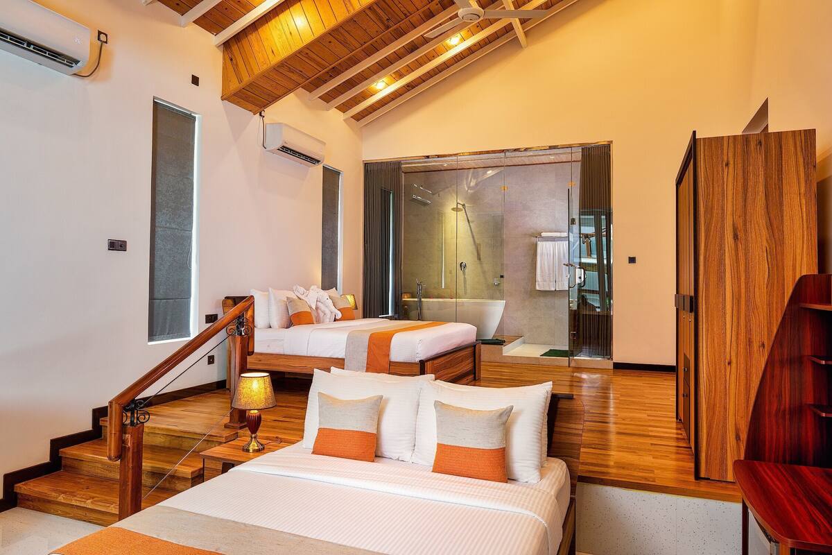 A hotel room with a wooden staircase, a large bed with white bedding and orange accents, a separate bathroom with a glass shower, and a wooden door.