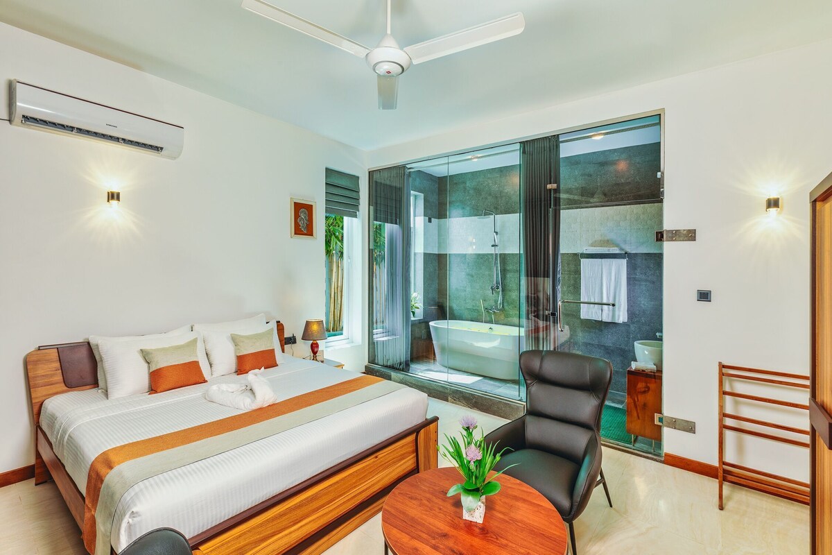 A modern hotel room with a large bed, a sitting area, a bathroom with a bathtub, and a view through a glass door.