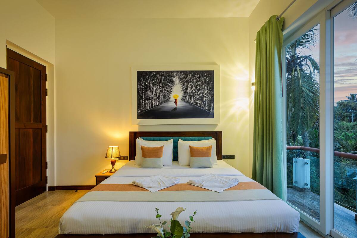 A hotel room with a large bed, a painting above the bed, a view of the outdoors through the window, and a wooden door.