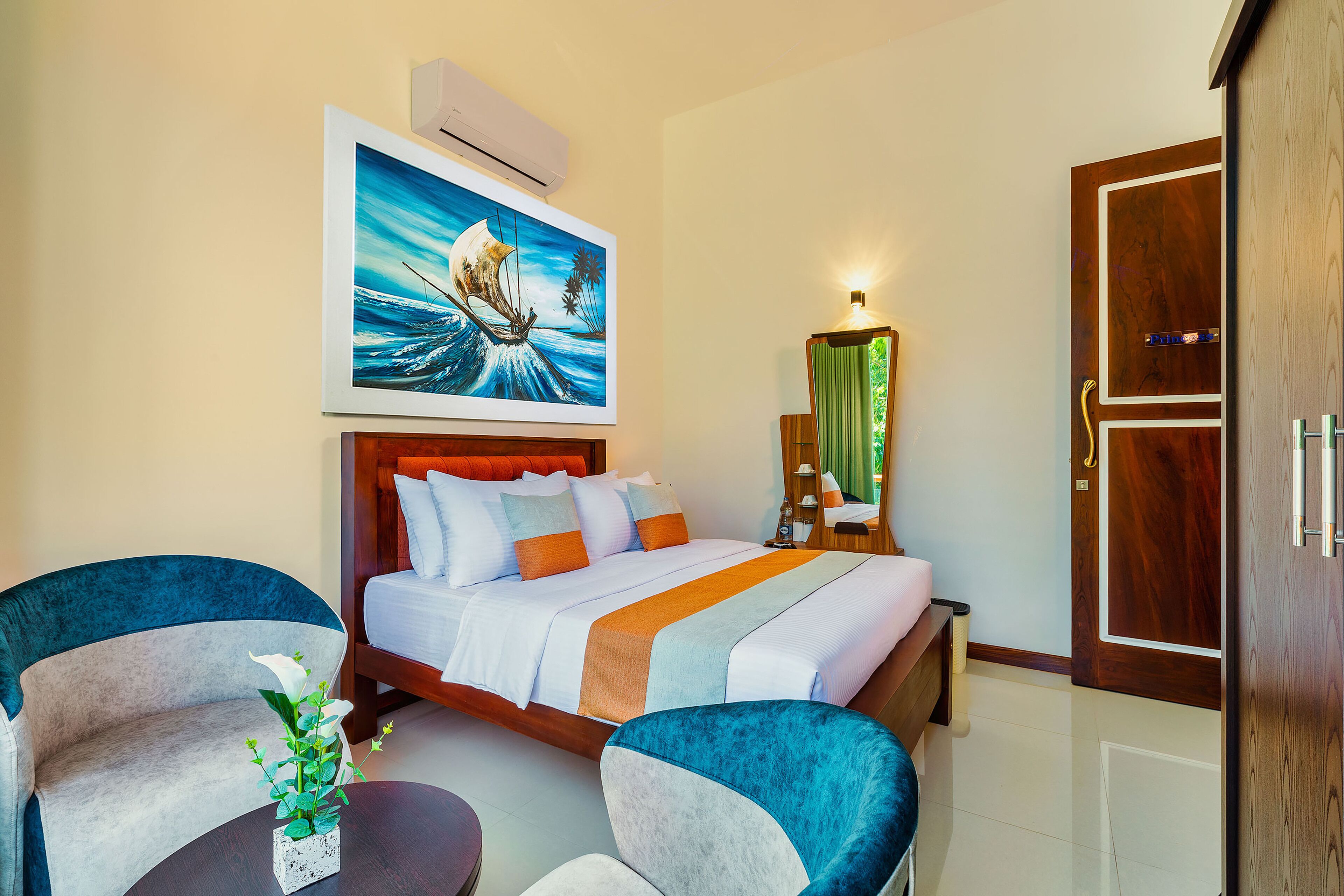 A hotel room with a bed, two armchairs, a small table with a vase, a painting above the bed, and a wooden wardrobe.