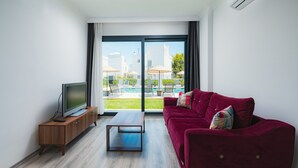 Ένα μοντέρνο σαλόνι με έναν κόκκινο καναπέ, μια τηλεόραση τοποθετημένη πάνω σε ένα ξύλινο έπιπλο και μια μεγάλη συρόμενη γυάλινη πόρτα που οδηγεί σε μια εξωτερική περιοχή με πισίνα.