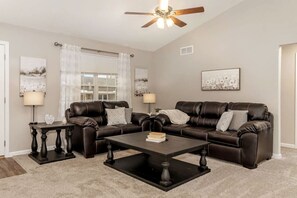 A living room with a dark brown leather sofa set, two side tables with lamps, a coffee table, a ceiling fan, and a window with curtains.