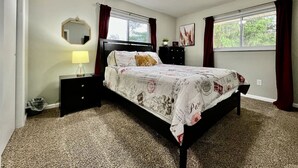 Un dormitorio con cama, mesitas de noche, lámpara, espejo, ventana con cortinas y un aplique de pared.