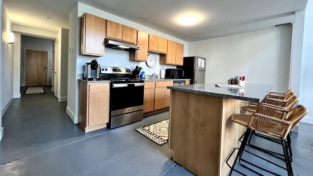 A modern kitchen with wooden cabinets, stainless steel appliances, and a central island with bar stools. - 5104 Cartridge Factory Loft (Maineville)