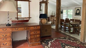 Una habitación con un escritorio de madera, cajones, una lámpara, un modelo de barco, un espejo, un armario, una alfombra y una zona de comedor con sillas y una mesa.
