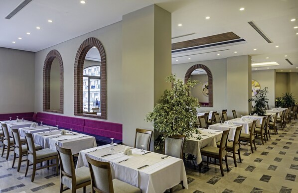 A hotel dining area with multiple tables set for guests, large windows with arched brick frames, and a central plant. - My Ella Bodrum Resort & Spa (Bodrum)