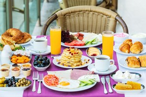 Un bufé de desayuno con diversos platos, como panqueques, huevos y pasteles, sobre una mesa con una silla de mimbre al fondo.