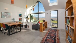 A spacious room with a large window, a dining area, a bookshelf, and a view of the outdoors. - The Brackens : Beachfront 3-Bed Estuary Home in Malltraeth | Managed by Anglesey Holiday Lettings (Malltraeth)