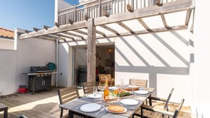 Un área de comedor exterior techada con mesa de madera para dos personas, una parrilla y un patio con piso de madera.