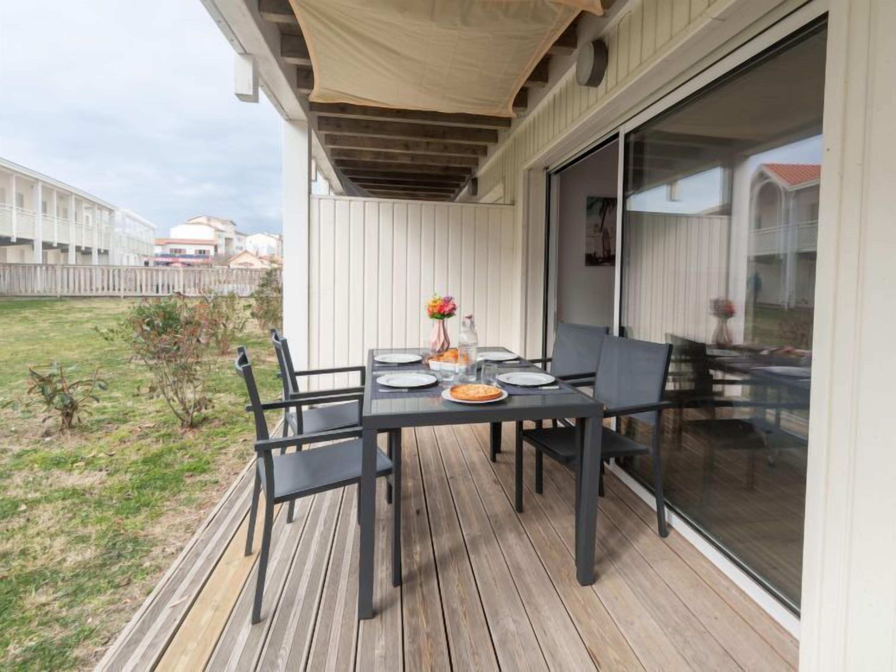 Un espacio cubierto para comer al aire libre con una mesa dispuesta para cuatro personas, rodeada de sillas, sobre una terraza de madera.