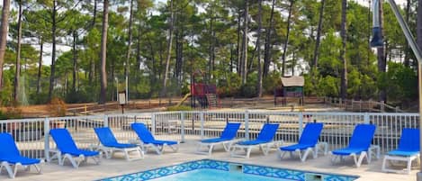 Piscina com espreguiçadeiras azuis e brancas, cercada por um muro branco e árvores ao fundo.
