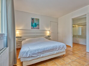 Quarto com cama, abajures, um quadro na parede e um banheiro visível através de uma porta aberta.