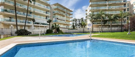 A swimming pool with a tiled deck, surrounded by multi-story buildings with balconies, palm trees, and a clear sky.