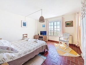 A bedroom with a bed, a desk with a TV, a chair, a window with a view, and a hanging light fixture.