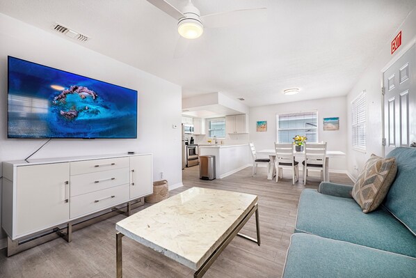 Una sala moderna con una televisión de pantalla plana, un sofá, una mesa de centro y una zona de comedor con una cocina al fondo.