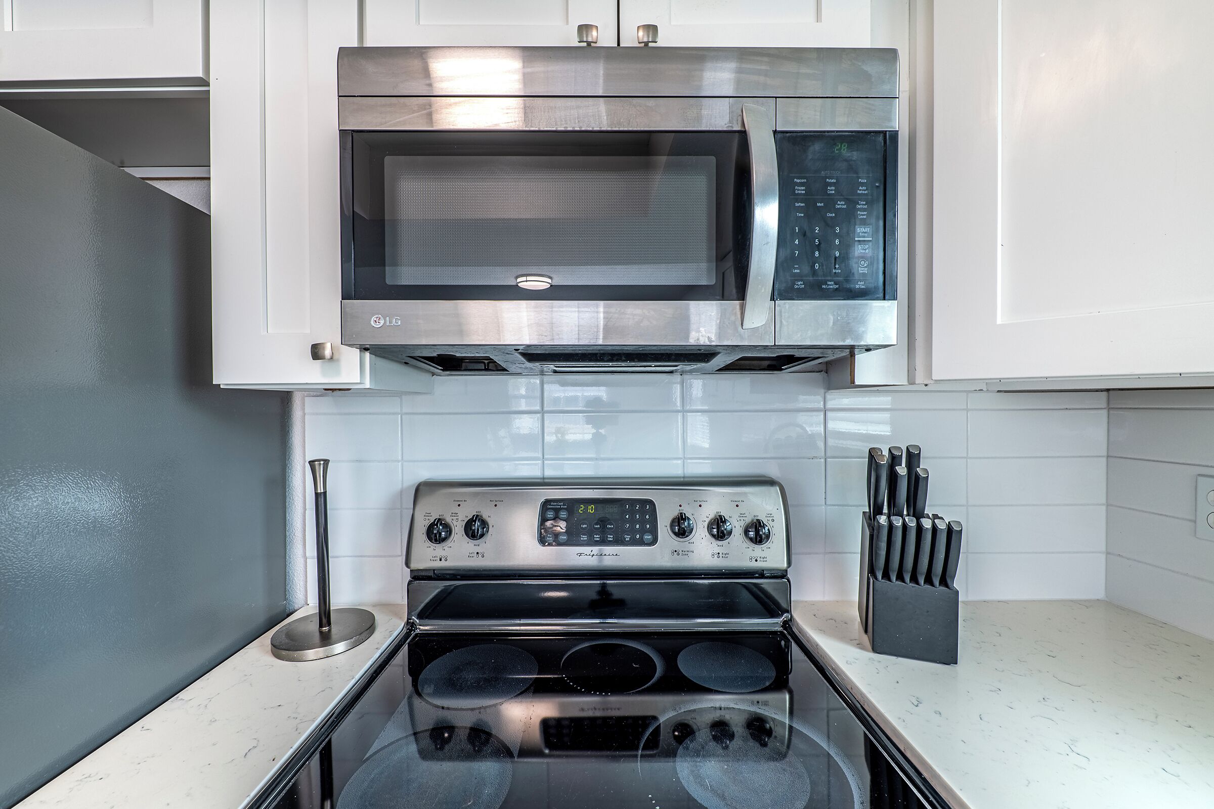Una cocina con microondas y horno de acero inoxidable, armarios blancos y un bloque de cuchillos sobre la encimera.