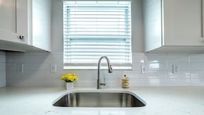 A kitchen with a stainless steel sink, white tiled backsplash, and a window with blinds. - Spacious 2-bed, 2-bath second-floor unit minutes from Indian Rocks Beach. Sleeps nine with full kitchen, WiFi, smart TVs, parking, beach gear, and direct pool access. A bright, comfortable base for families and groups (Indian Rocks Beach)