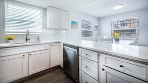 A modern kitchen with white cabinetry, a dishwasher, and a window with blinds. - Spacious 2-bed, 2-bath second-floor unit minutes from Indian Rocks Beach. Sleeps nine with full kitchen, WiFi, smart TVs, parking, beach gear, and direct pool access. A bright, comfortable base for families and groups (Indian Rocks Beach)
