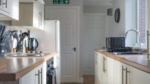 A compact kitchen with white cabinets, a wooden countertop, a microwave, a kettle, and a fire exit sign. - Lily Apartment 2 - 2 bed flat Bedlington (Bedlington)