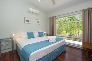A bedroom with a large bed, a nightstand with a lamp, an air conditioner, and a window with curtains. - Shipwreck Sands (Picnic Bay)