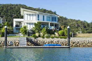 Una casa de dos pisos con un muelle, rodeada por una costa rocosa y abundante vegetación.