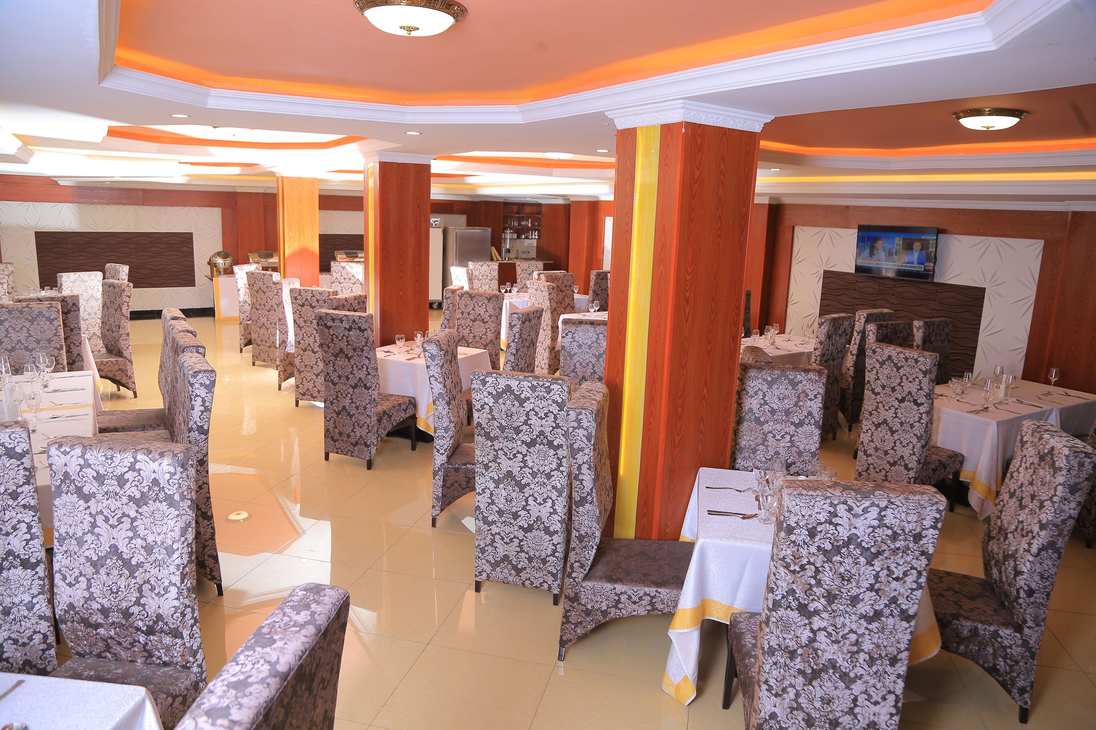 A banquet hall with rows of floral-patterned chairs and tables, a central column, and a television screen on the wall.