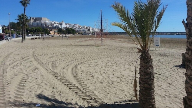 Een zandstrand met voetafdrukken, palmbomen en een gebouw in de verte.