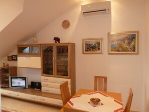 A dining area with a wooden table, chairs, and a TV displaying a nature scene. There is a cabinet with glass doors containing glasses and a small elephant figurine on top. Two framed paintings are on the wall.