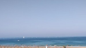 Una playa con cielo despejado, mar tranquilo y algunas embarcaciones.
